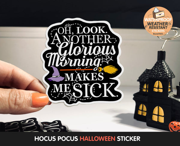 Hand holding a Oh Look Another Glorious Morning Makes Me Sick sticker against a spooky table scene. The sticker features a smooth black background with a purple witches hat, orange and gold witches broom, and modern white text