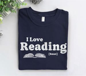 May include: A navy blue t-shirt with white text that reads &#39;I Love Reading (Smut)&#39; and a white outline of an open book.