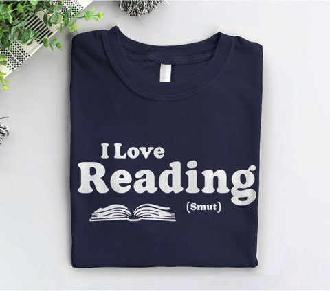 May include: A navy blue t-shirt with white text that reads &#39;I Love Reading (Smut)&#39; and a white outline of an open book.